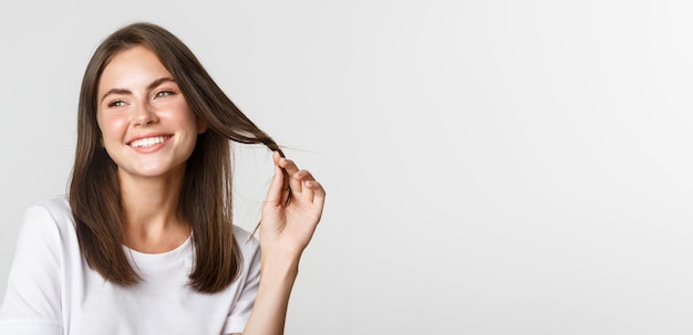 Foto gratuita ritratto di bella ragazza civettuola che gioca con i capelli e sorride alla telecamera bianca in piedi civettuola