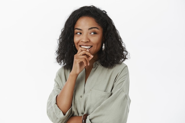 Portrait of flirty and curious feminine cute dark-skinned female coworker in blouse holding finger on lip and smiling with desire