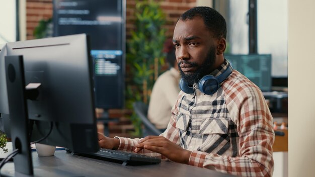 Portrait of focused african american programer wearing wireless headphones working looking at computer screen while typing. Software developer writing code in it agency office.