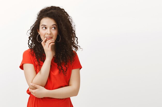 Portrait of focused dreamy woman with curly hair in fashionable casual red dress, holding hand on chin