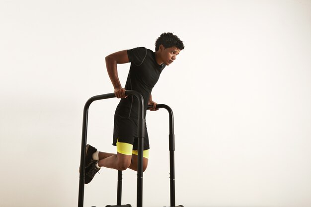 A portrait of a focused muscular African American young man in black workout clothes doing dips on parallel bars on white