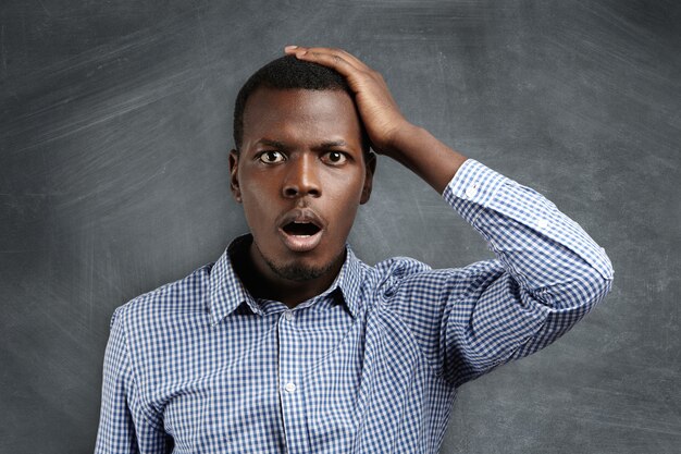 Portrait of forgetful young dark-skinned customer or employee wearing checkered shirt looking with shocked and guilty expression, holding hand on his head, opening mouth widely.