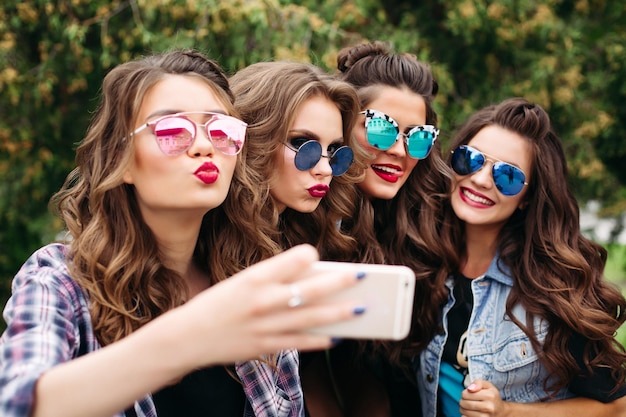 Portrait of four girlfriends wearing casual trendy clothes, hairstyles and make up posing while making self-portrait via cell phone sitting outside in park on day.