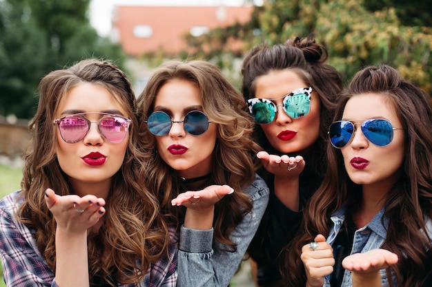 Portrait of four girlfriends wearing casual trendy clothes, hairstyles and make up posing while making self-portrait via cell phone sitting outside in park on day.