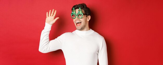 Portrait of friendly cheerful man in party glasses and white sweater saying hello and looking left g...