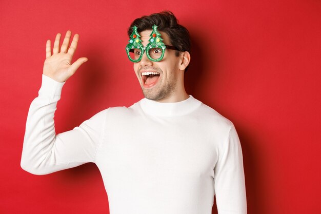 Portrait of friendly, cheerful man in party glasses and white sweater, saying hello and looking left, greeting a friend, standing over red background