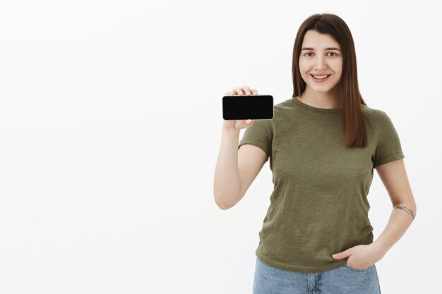 Portrait of friendly and happy young pleased positive brunette in casual t-shirt smiling and showing smartphone in horizontal position as presenting app or mobile phone over gray wall