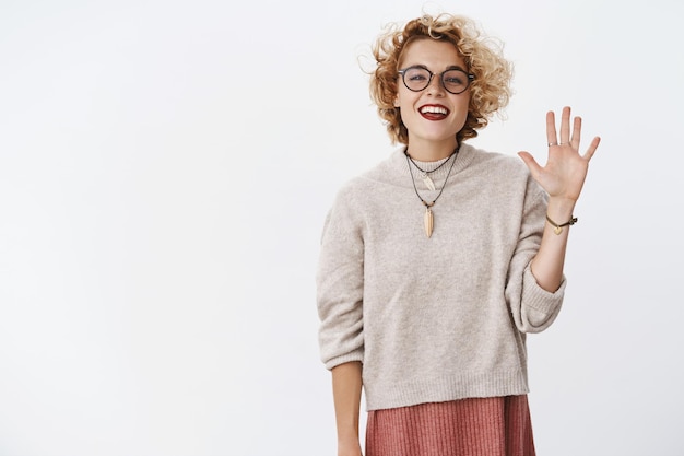 Portrait of friendly-looking outgoing and relaxed carefree attractive hipster girl with glasses and sweater raising palm to wave hi and greet new members of group smiling joyfully