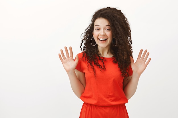 Portrait of friendly-looking stylish european woman with curly hair in red dress, raising palms from unexpected meeting with friend