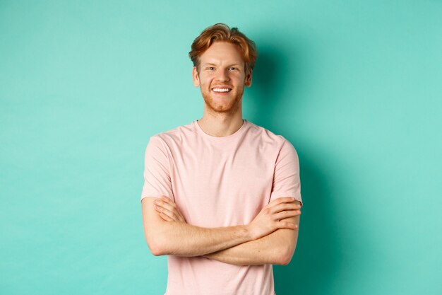Portrait of friendly-looking young man with red hair and beard smiling and starng satisfied, holding hands crossed on chest, standing over turquoise background