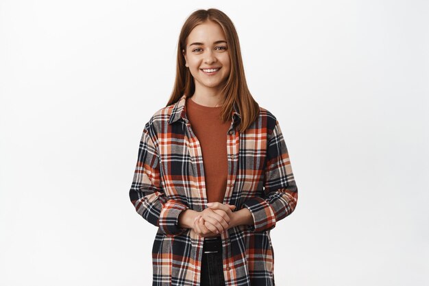 Portrait of friendly and polite caucasian woman, helpful woman assistant smiling pleasant, holding hands together clenched over chest, standing against white wall