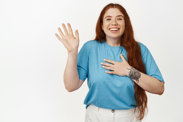 Portrait of friendly redhead girl, waving hand with cheerful face, introduce herself, tell her name, greeting someone, standing in t-shirt on white.