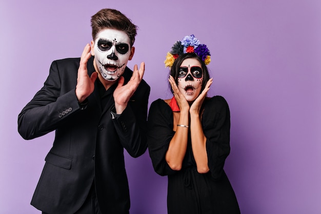 Portrait of frightened and shocked boy and girl. Couple with face art on day of all dead in dark attires posing on lilac wall.