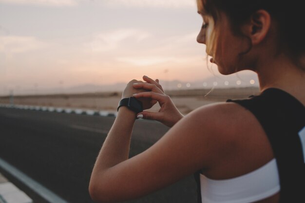 道路上の手で時計を見てスポーツウェアの後ろから美しい若い女性の肖像画。晴れた夏の早朝、ファッショナブルなスポーツウーマンのトレーニング、モチベーション