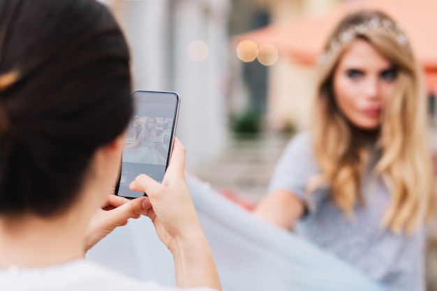 Портрет сзади брюнетка девушка, делающая фото на телефон довольно белокурой женщины на улице.