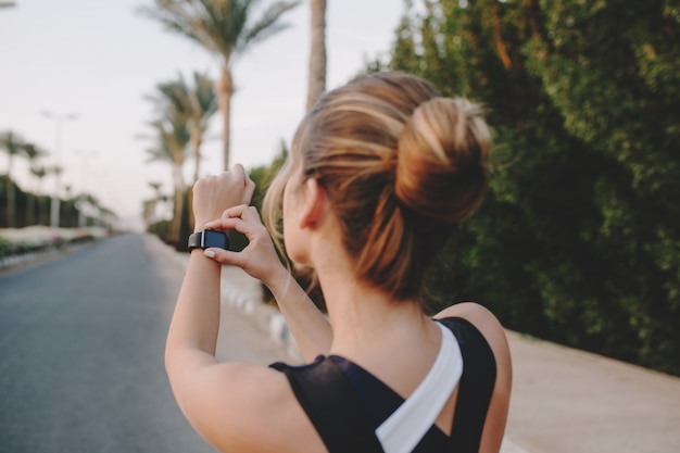 Портрет со спины модной спортсменки, смотрящей на современные часы на руках на улице с пальмами тропического города. Тренировка привлекательной женщины, тренировки, здоровый образ жизни, трудолюбие