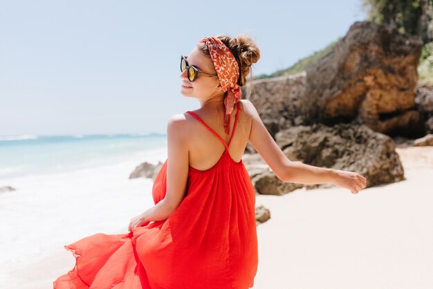 夏の暖かい日に人生を楽しんでいる嬉しい白人の女の子の後ろからの肖像画。ビーチで踊る赤いドレスを着たヨーロッパの魅惑的な女性の屋外写真