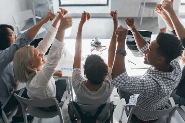 Free photo portrait from back of glad students sitting together at the table and raising hands. indoor photo of team of freelance specialists having fun after hard work in office.