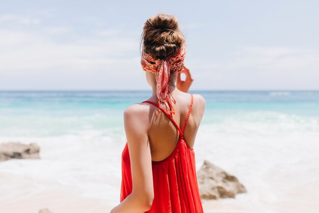로맨틱 젊은 아가씨 뒤에서 초상화는 해변에서 휴식하는 동안 빨간 옷차림을 착용합니다. 오션 뷰를 즐기는 행복한 여자의 야외 샷.
