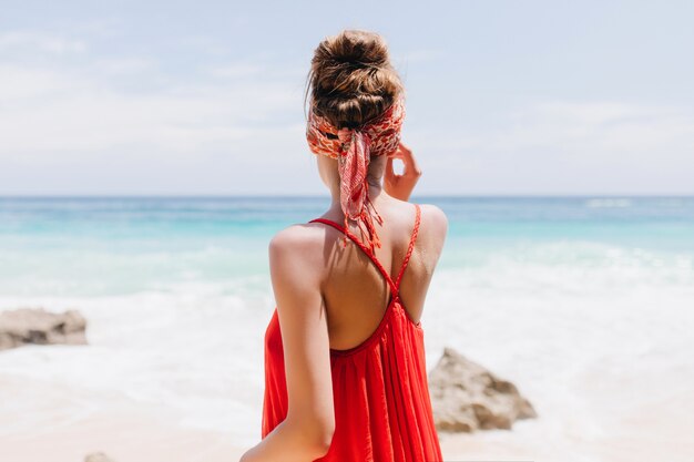 Portrait from back of romantic young lady wears red attire during rest at beach. Outdoor shot of blissful girl enjoying ocean view.