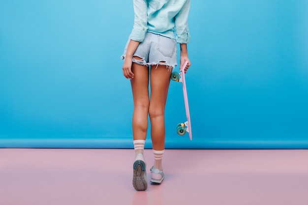 Portrait from back of sporty tanned woman wears cute socks and cotton shirt. Indoor photo of graceful girl with bronze skin in denim shorts holding longboard.