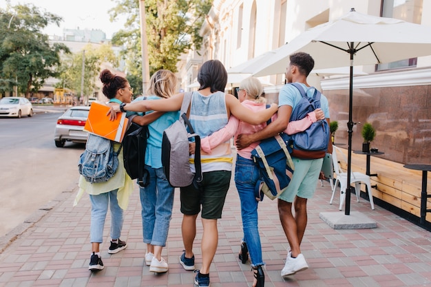 Foto gratuita ritratto dal retro di studenti con eleganti zaini che camminano per strada dopo le lezioni all'università. un giovane uomo alto e castano che abbraccia le ragazze mentre trascorre del tempo con loro da superare.