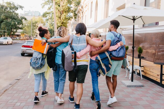 Portrait from back of students with stylish backpacks walking down the street after lectures in university. Tall brunette young man embracing girls while spending time with them outdoot..
