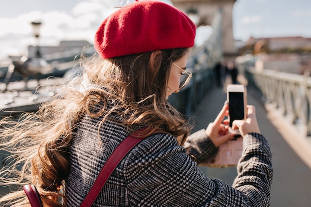 Portrait from back of stylish woman in tweed jacket holding phone and making photo of city landscape