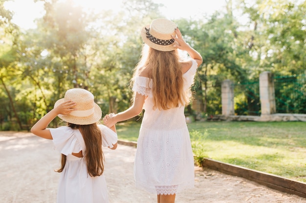 Portrait from back of tall tanned woman leading daughter down the street. Blonde slim lady holding hands with little brunette girl, walking by lawn and fence in park.