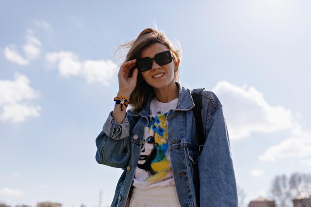 サングラスとデニムのジャケットと青と黄色のプリントのtシャツで笑顔の幸せな女の子の下からの肖像画は、メガネに触れて、青い空を背景にカメラに微笑んでいます