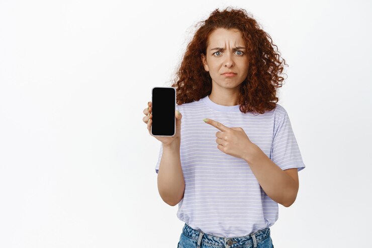Portrait of frowning disappointed woman pointing at mobile phone screen. Attractive young female model showing smth concerning, disappointing app on smartphone.