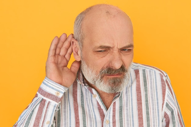 Free photo portrait of frowning frustrated bearded male pensioner in striped shirt keeping hand at his ear, listening attentively, trying to hear indistinct talking. hearing problems and eavesdropping