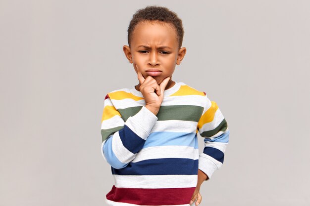 Portrait of frowning grumpy little dark skinned boy expressing unwillingness or disagreement. Serious African child in stylish jumper holding hand on his chin, having frustrated pensive look