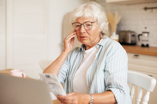 Free photo portrait of frustrated gray haired female pensioner wearing eyeglasses sitting at kitchen table with laptop, holding bill and touching face, shocked with amount of total sum for electricity