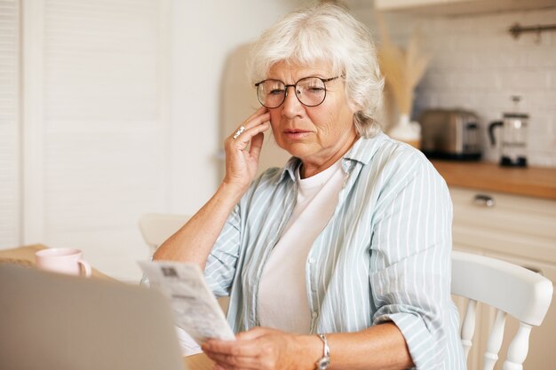 Portrait of frustrated gray haired female pensioner wearing eyeglasses sitting at kitchen table with laptop, holding bill and touching face, shocked with amount of total sum for electricity