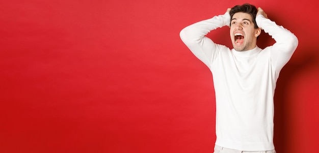 Portrait of frustrated man in white sweater, screaming in panic and looking at upper left corner, have terrible problem, standing over red background.