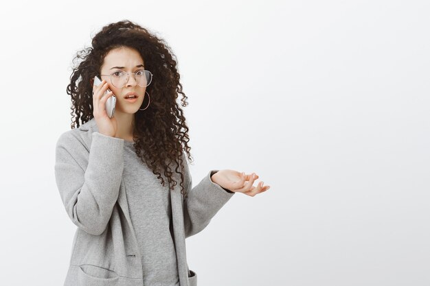 Portrait of frustrated questioned girlfriend with curly hair in grey coat and glasses, raising hand cluelessly while talking on smartphone