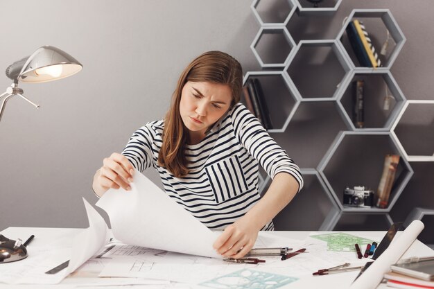 Portrait of frustrated young european dark-haired freelance manager talking on phone with team leader, trying to find mistake in project blueprints. Freelance, teamwork concept