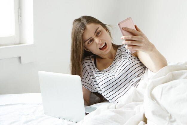 彼女はオンライン支払いを行うことができないか、接続に問題があるので、開いたラップトップでベッドに横たわって、携帯電話を持って怒って叫んでいる欲求不満の若い女性フリーランサーの肖像画