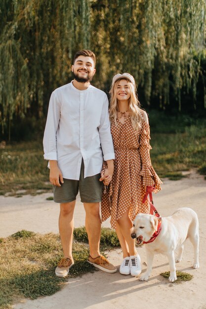 Portrait full-length of married couple posing with their white Labrador in park against background of willow.