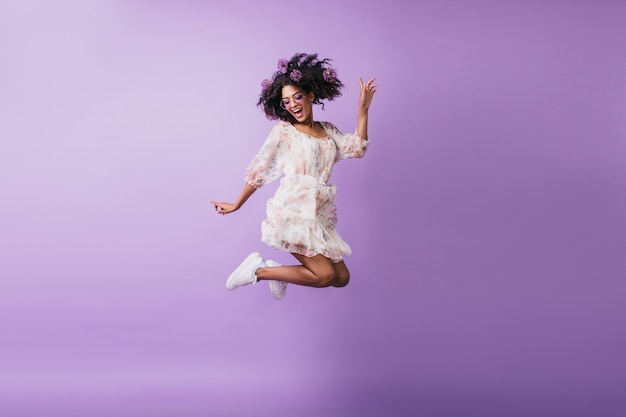 ジャンプする白い服装で面白いアフリカの女の子の肖像画。ポジティブな感情を表現する明るいブルネットの若い女性。