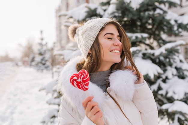 Portrait funny amazing woman enjoying winter time, holding lollypop on street. Brightful happy emotions of young woman in warm white winter clothes with closed eyes, great smile.