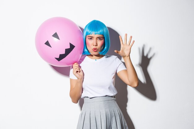 Portrait of funny asian girl trying to scare someone on halloween, wearing blue wig and holding pink balloon with scary face.