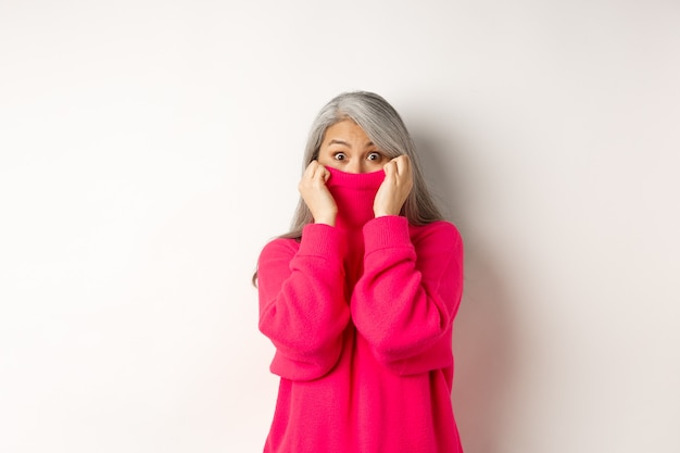 Free photo portrait of funny asian grandmother hiding face in sweater collar, peeking at camera silly, standing over white background