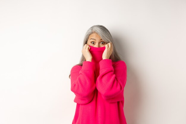 Портрет смешной азиатской бабушки, пряча лицо в воротнике свитера, глупо глядя в камеру, стоя на белом фоне