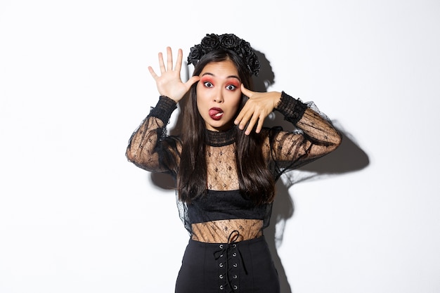 Portrait of funny asian woman in witch costume celebrating halloween, mocking someone, showing faces and sticking tongue, standing over white background.