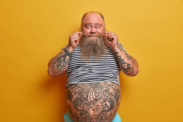 Free photo portrait of funny bearded man curls mustache, has tattooed arms and tummy, dressed in undersized striped sleevless t shirt, has problem of obesity and overweight, isolated on yellow wall