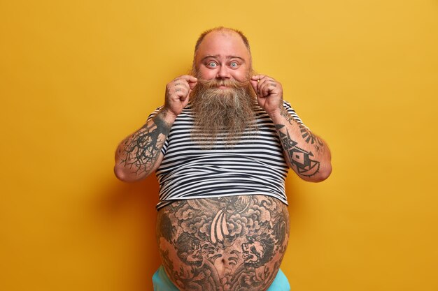 Portrait of funny bearded man curls mustache, has tattooed arms and tummy, dressed in undersized striped sleevless t shirt, has problem of obesity and overweight, isolated on yellow wall