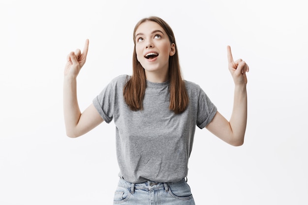 Portrait of funny beautiful european girl with dark hair with grey t-shirt and jeans looking upside with dreamy face expression, pointing on white wall with fingers on both hands. Copy space