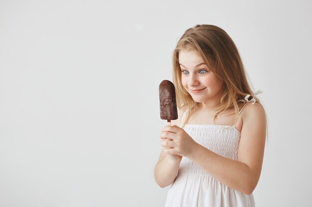 Portrait of funny blonde little hair in white dress looking at ice-cream in her hands with happy and excited look, going to eat it.